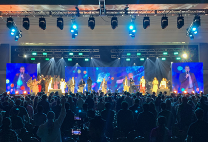 Large screens on stage with performers in front and on screens, with large audience in front