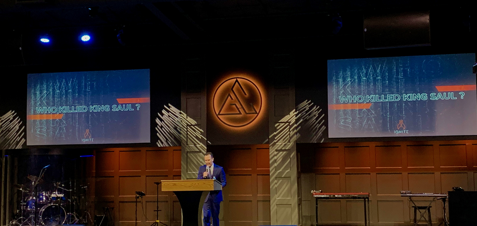 Pastor preaching on stage with church logo on wall behind him and two large screens on both sides of him